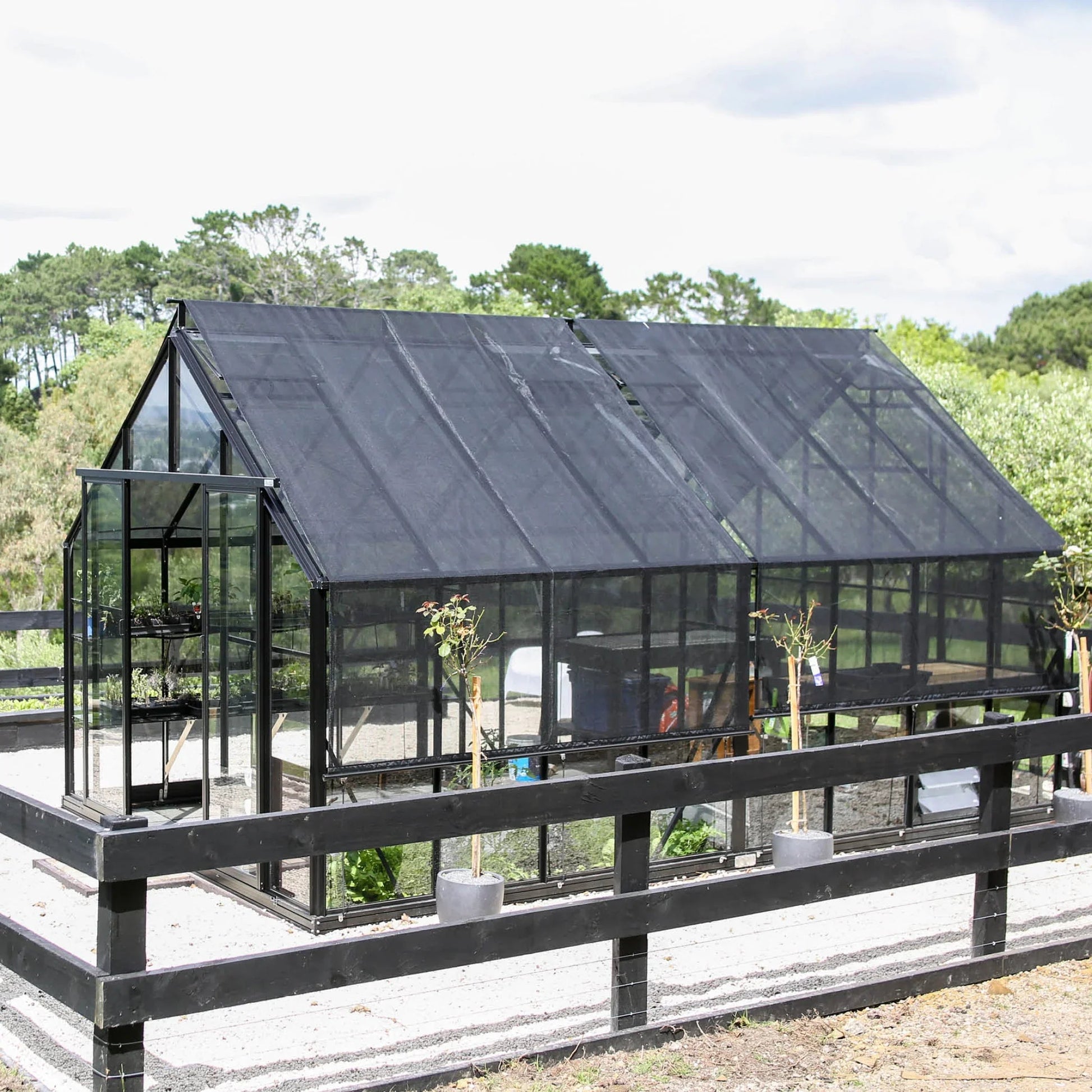 Black metal framed greenhouse with black shade cover roof panels surrounded by black fence and potted plants