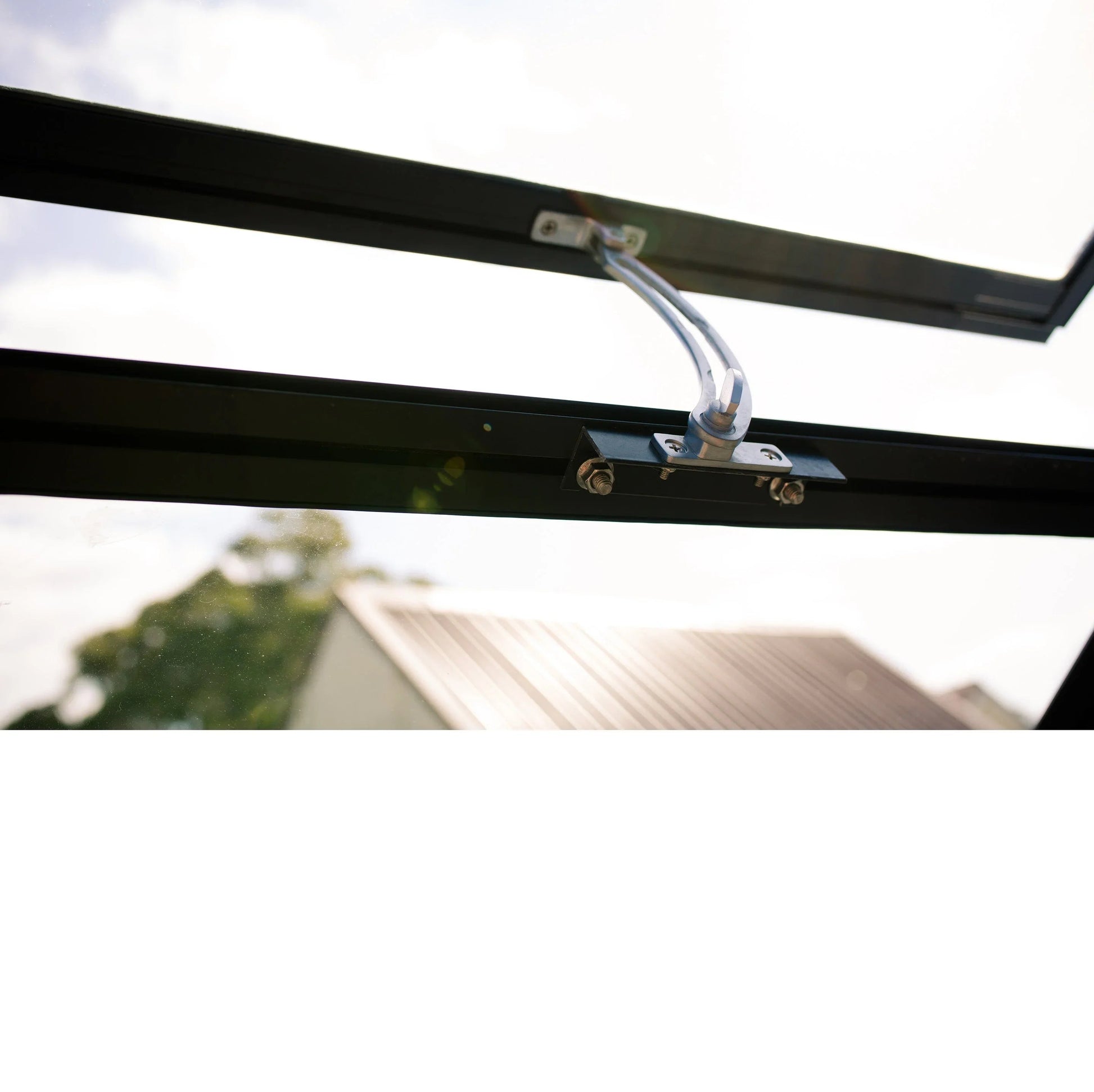 Close-up of a black metal window vent opener with mounting screws against a bright sky background